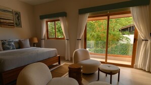 A bedroom with a bed, two chairs, a round wooden stool, and a window with curtains.