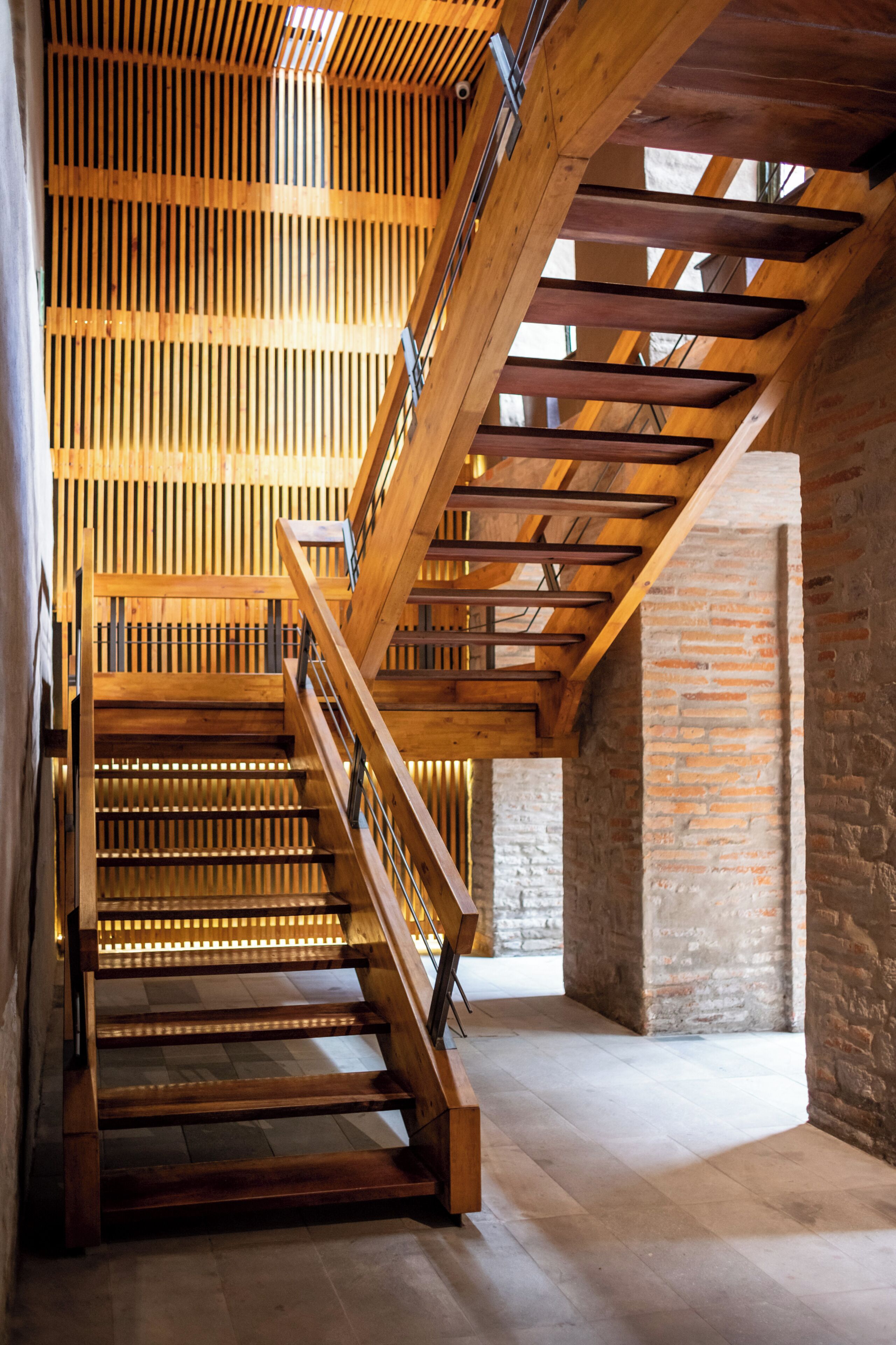 Una escalera de madera con barandilla metálica que sube hacia un techo alto con listones de madera.