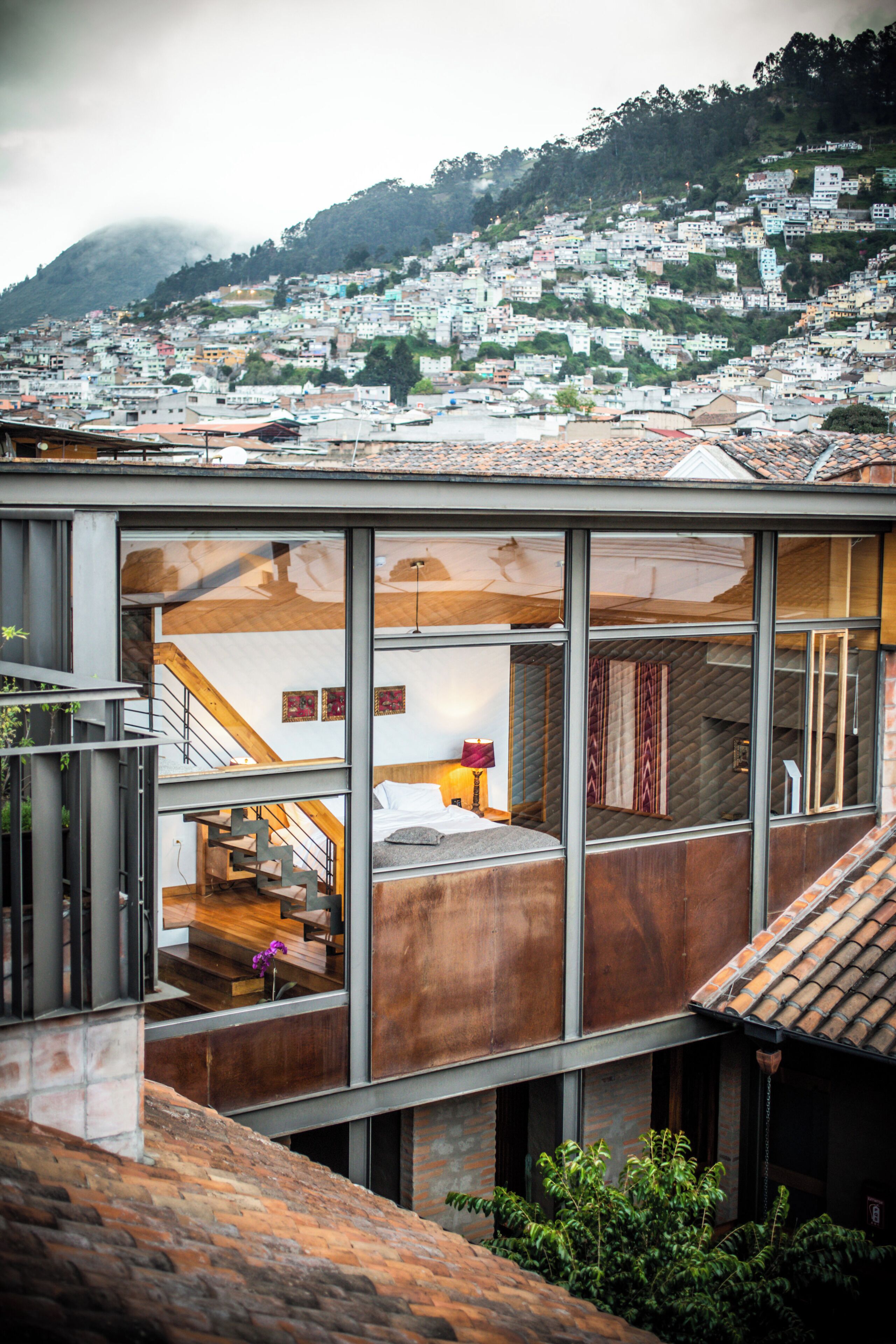 Un edificio moderno con fachada de vidrio, un interior visible con cama y escalera, y vista a un pueblo en la ladera.