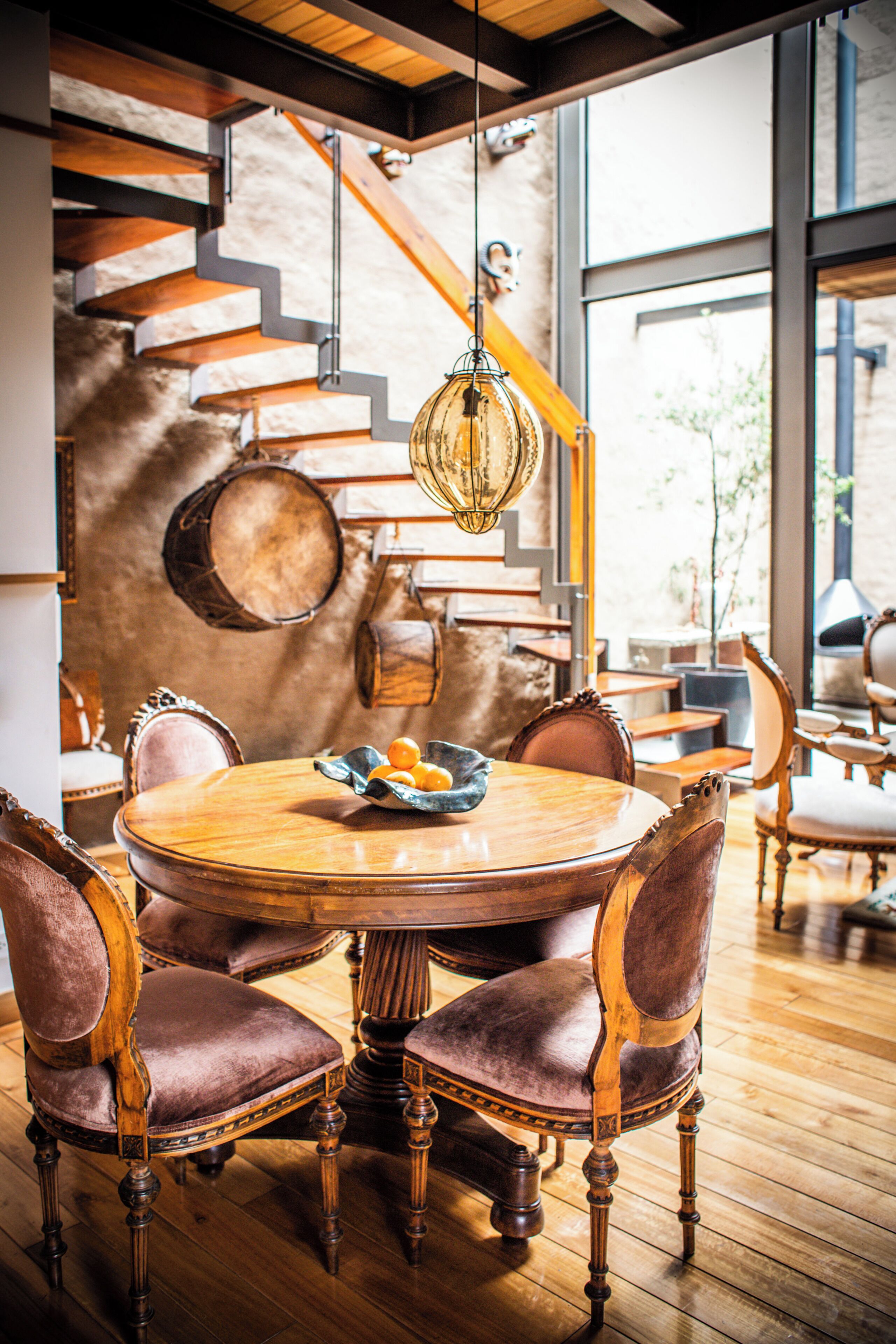 Un comedor con mesa y sillas de madera, una lámpara colgante y una escalera de madera con barandilla metálica.