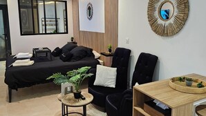 A modern living room with a bed, a black sofa, a round wooden side table, and a small potted plant.