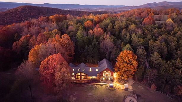 Une grande cabane en rondins à plusieurs étages, dotée d’une véranda qui fait le tour de la maison, et entourée d’une forêt dense aux couleurs automnales.
