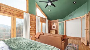 A bedroom with a large bed, a sofa, a ceiling fan, wooden walls, and a view through a window. - Luxury Shenandoah Cabin w/ Views + Hot Tub + Sauna (Luray)