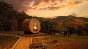 Une structure en bois en forme de tonneau, dotée d’une porte, entourée d’une terrasse et de chaises, le tout se dressant sur un fond de paysage montagneux aux couleurs automnales.