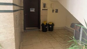 Un pasillo estrecho con una puerta de madera oscura, dos botes de basura negros y un suelo de baldosas.
