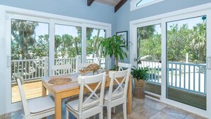 Un comedor con una mesa de madera y sillas blancas, ventanas amplias con vista a palmeras y un techo con vigas a la vista.