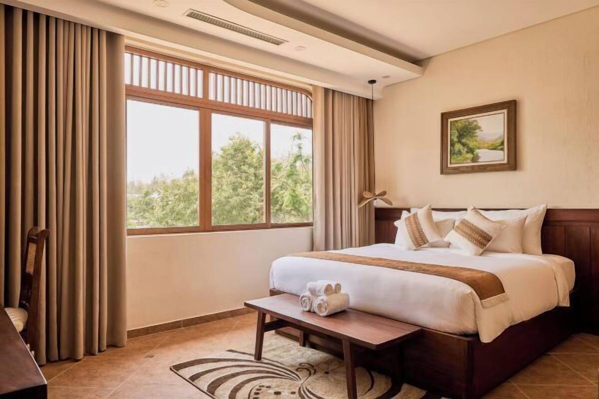 A hotel room with a bed, a desk, a chair, a window with curtains, and a painting on the wall.