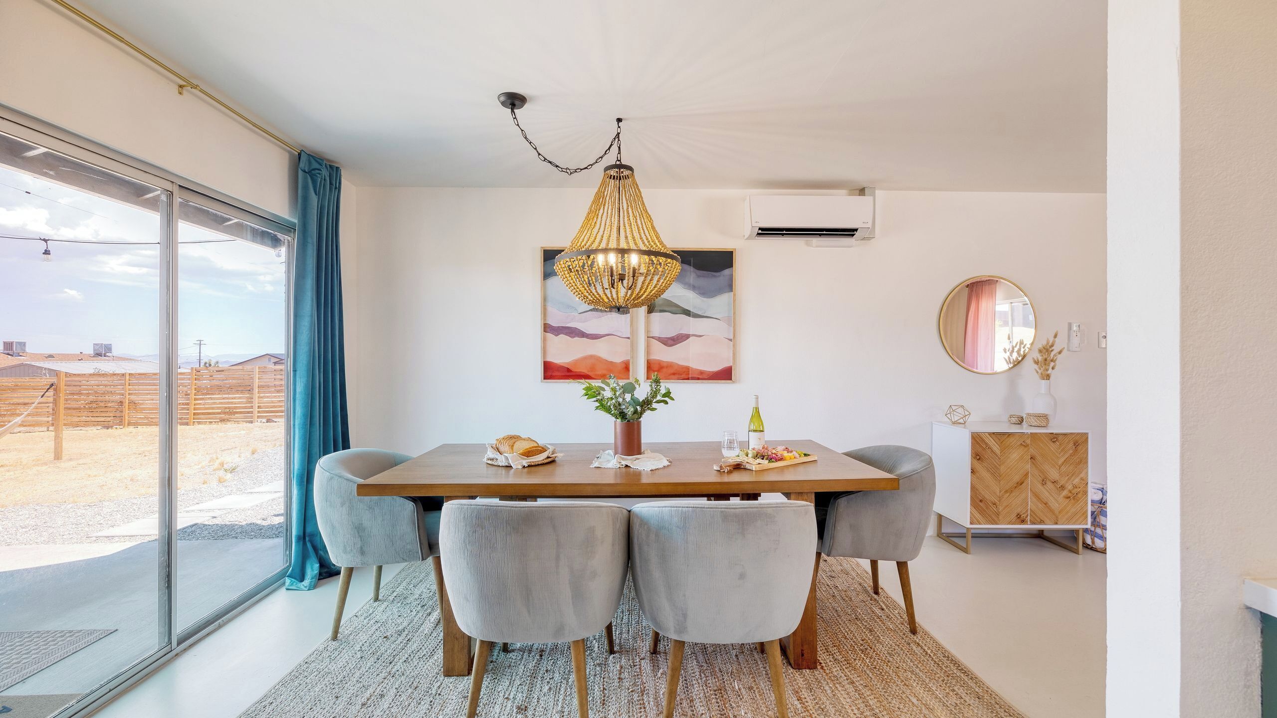 A dining area with a wooden table, grey upholstered chairs, a woven chandelier, and a large window with a view of a fenced outdoor area.