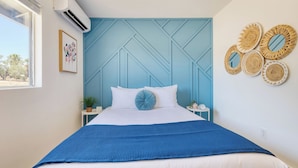 Een slaapkamer met een blauw hoofdeinde, witte beddengoed, een blauwe plaids, een potplant en wanddecoratie van geweven materiaal.