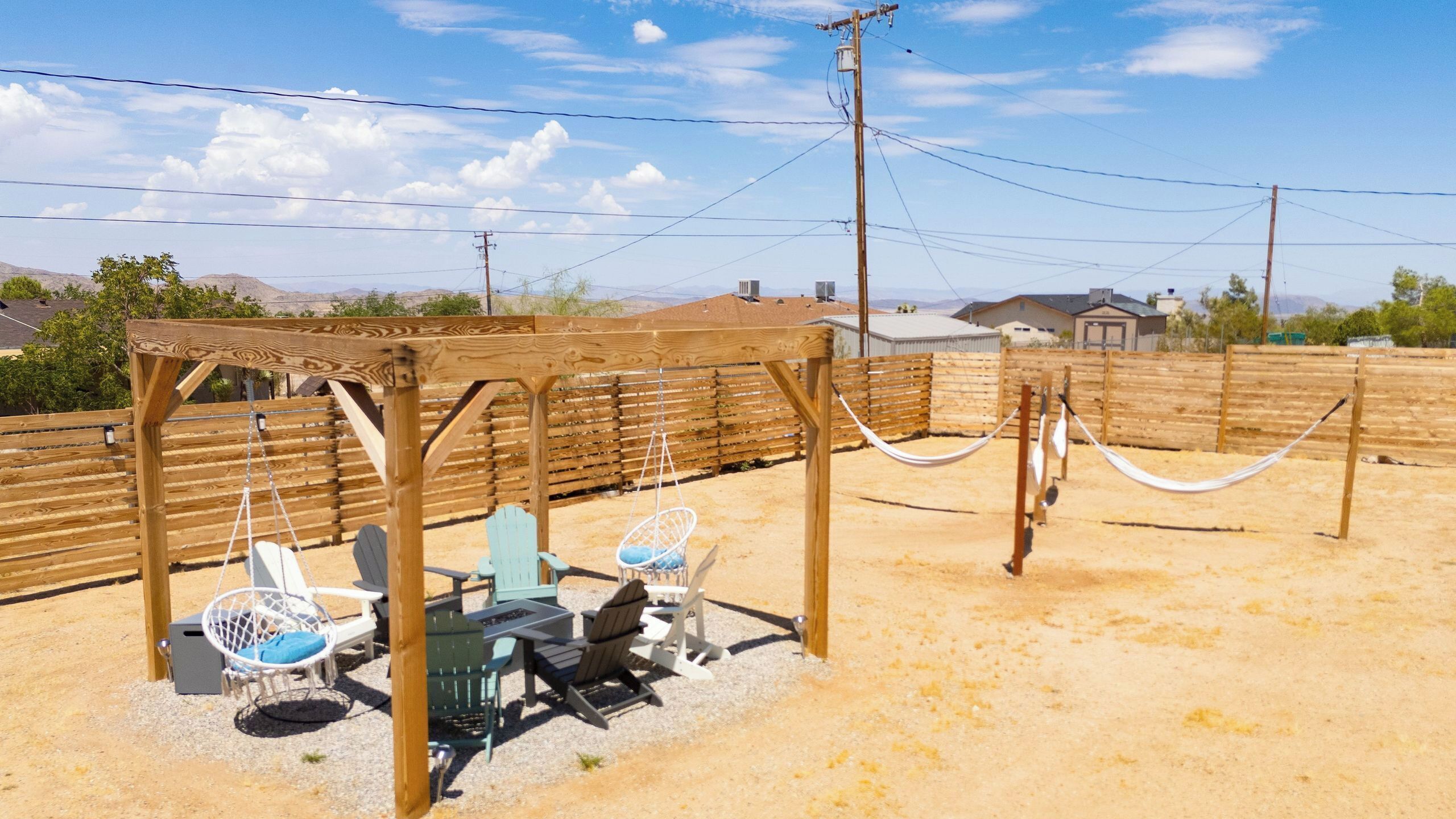 A wooden pergola with seating, including chairs and a hammock, in a desert setting with houses in the background.