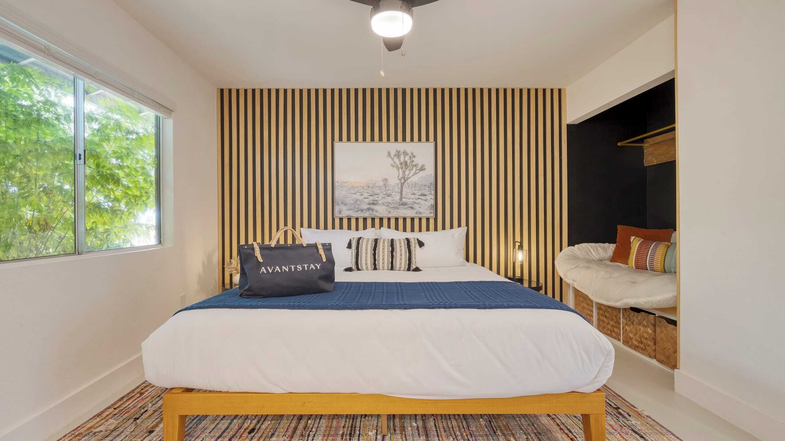 A bedroom with a bed, a nightstand, a chair, a window, and a wall with vertical stripes.