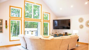 A modern living room with a large sectional sofa, a flat-screen TV, and large windows with a view of trees.