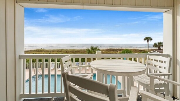 Un balcón con vista a la playa, una piscina y muebles blancos.