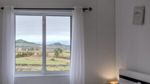 Ein Zimmer mit einem großen Fenster, das einen Blick auf eine ländliche Landschaft bietet, darunter Felder und ferne Hügel.