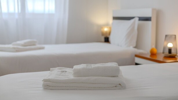 Ein Hotelzimmer mit zwei Betten, jedes mit einem ordentlich gefalteten weißen Handtuch. Nachtische mit Lampen und ein kleiner Tisch mit einer Lampe stehen bereit.