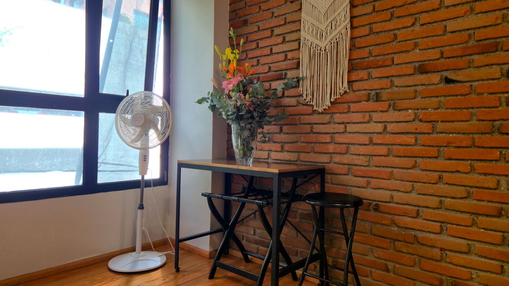 Una habitación con pared de ladrillo, un ventanal grande, un ventilador de pie, una mesita con un jarrón de flores y un taburete.