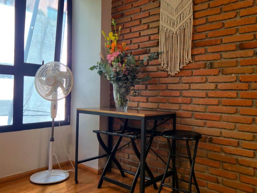 Una habitación con pared de ladrillo, un ventanal grande, un ventilador de pie, una mesita con un jarrón de flores y un taburete.