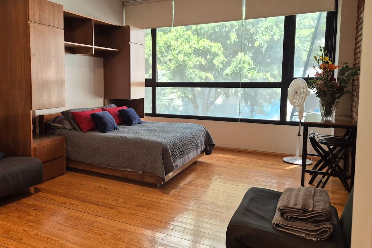 A bedroom with a bed, a sofa, a desk, and a window with a view of trees.