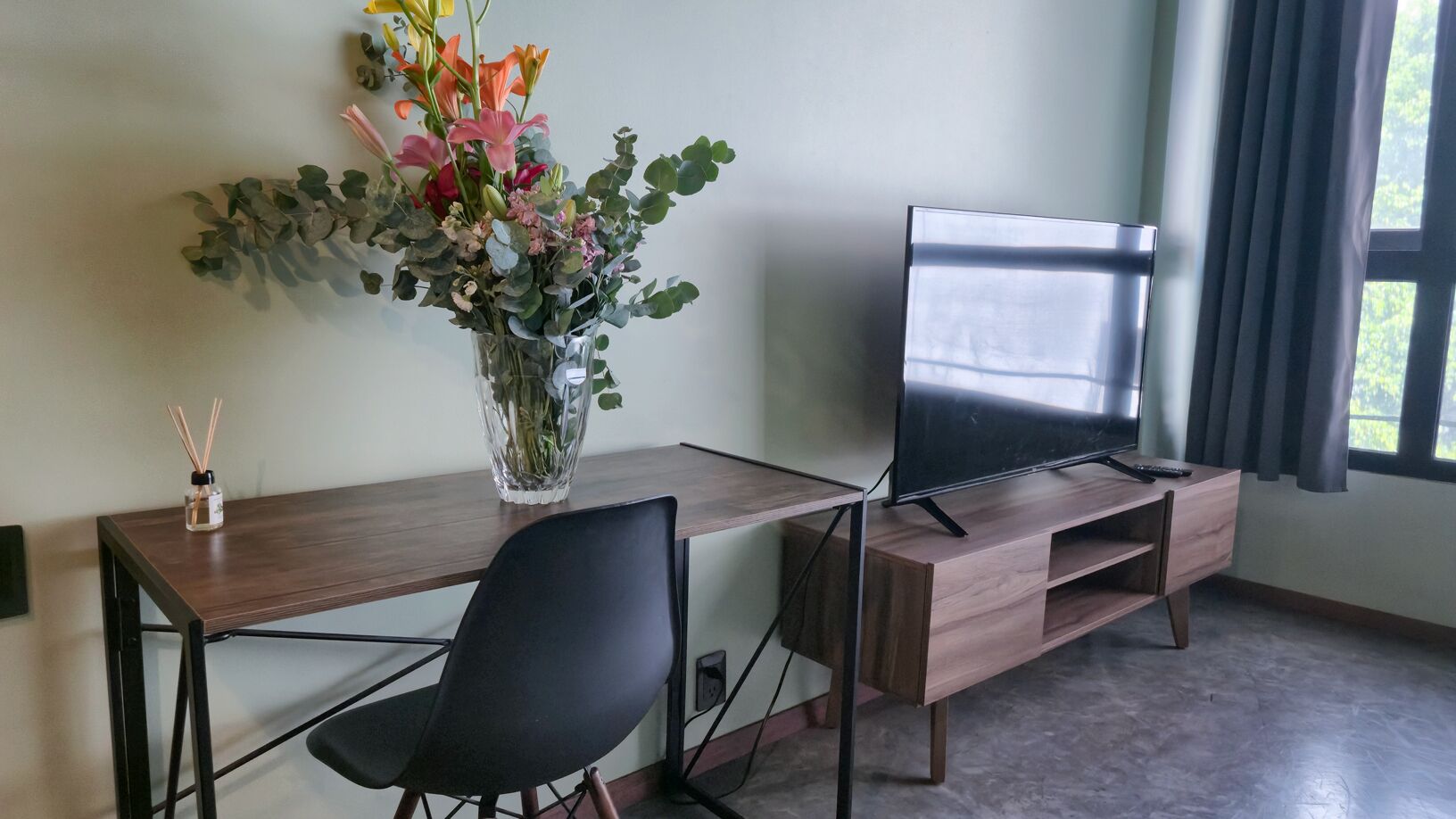Une chambre moderne avec un bureau en bois, une chaise de bureau noire, un téléviseur posé sur un meuble en bois et un vase avec des fleurs sur le bureau.