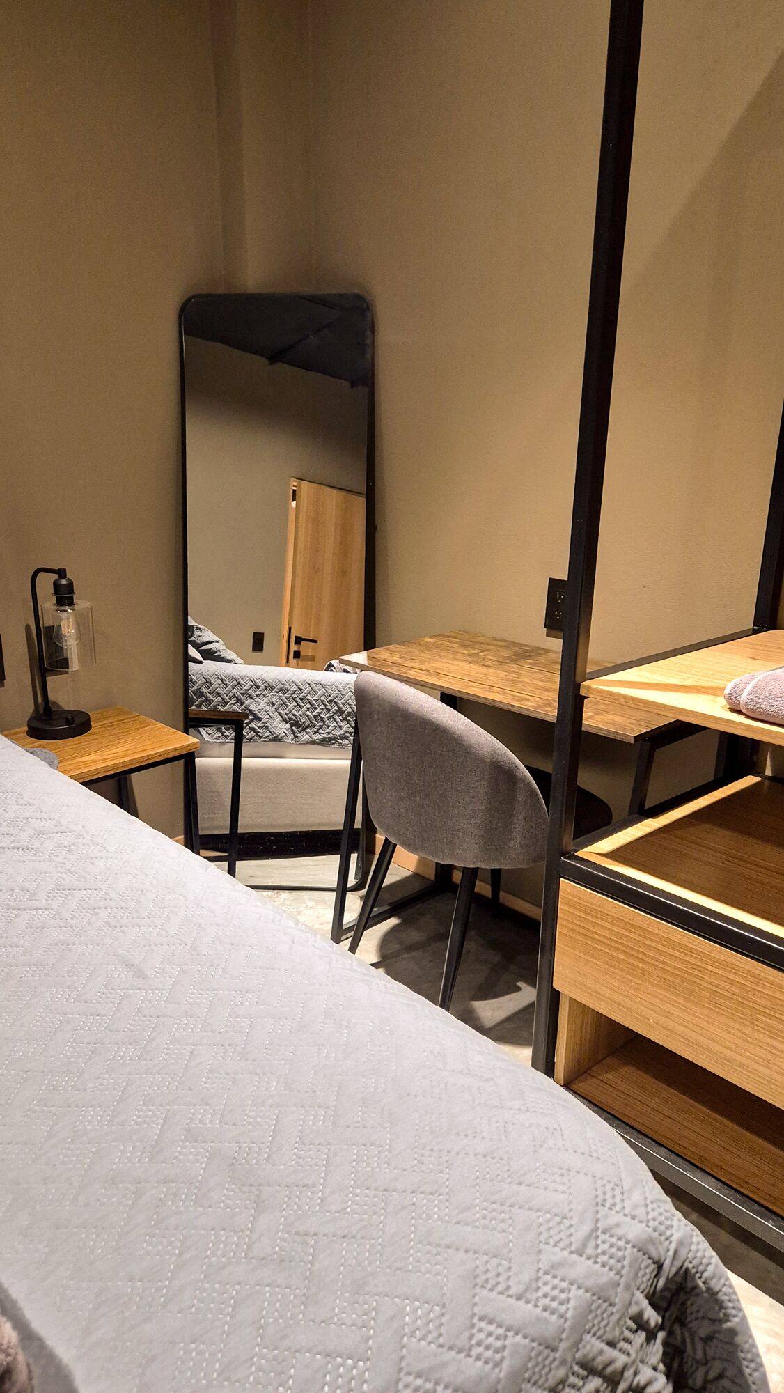 A compact room with a bed, a desk, a chair, and a mirror.