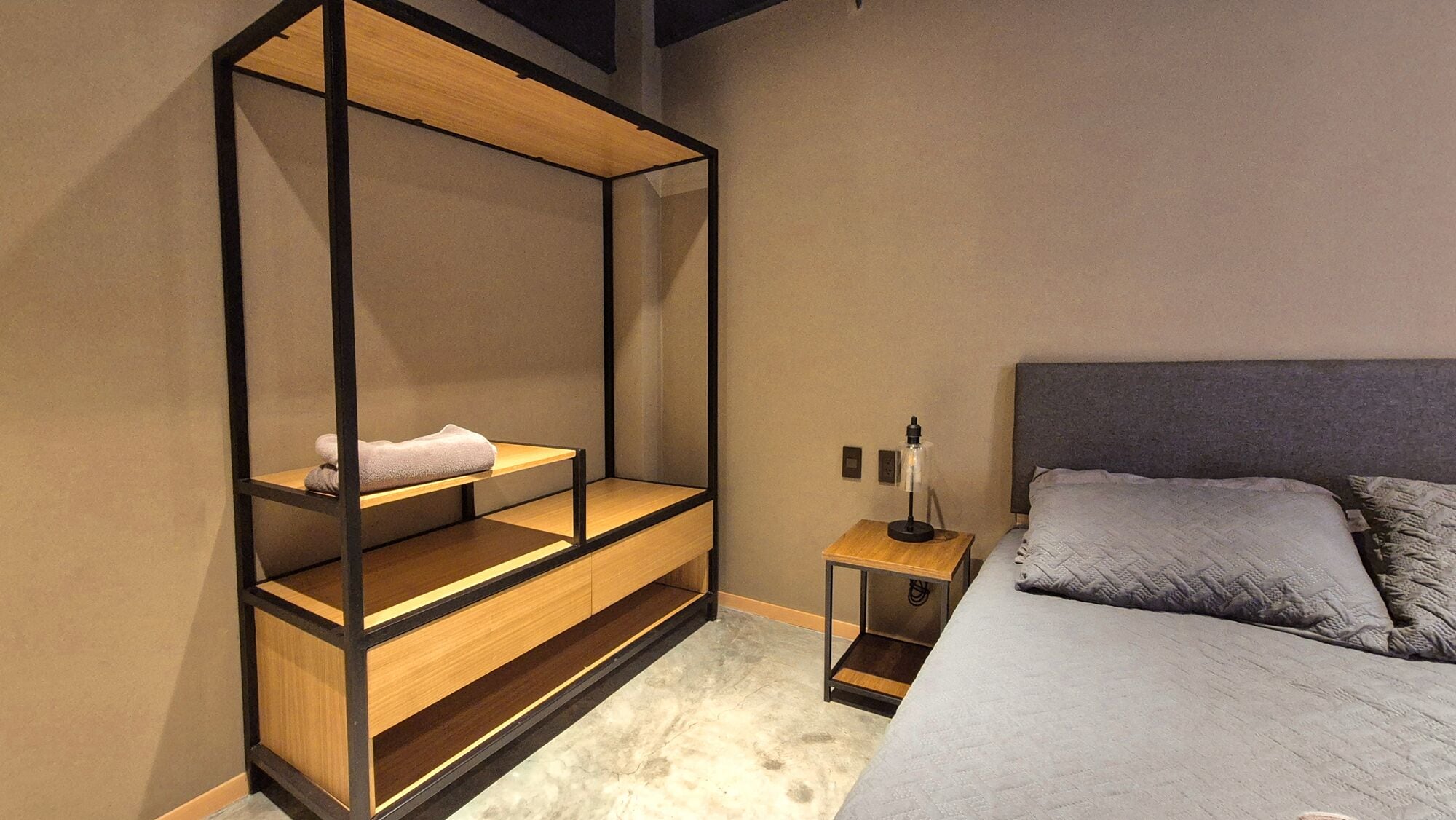 A bedroom with a bed, a nightstand, a wooden shelving unit, and a wall-mounted shelf.