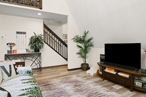 Ein Wohnzimmer mit einer Couch, einem Fernseher, einem Couchtisch, einer Treppe und zwei Blumentöpfen.