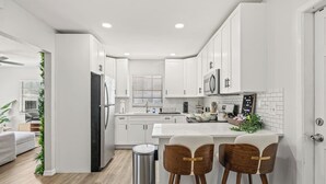 A modern kitchen with white cabinets, a stainless steel refrigerator, a microwave, and a sink. There are two wooden bar stools and a trash can. - 4BR Oasis | Jacuzzi/Gym & Game room | Sleeps 12! (Tampa)