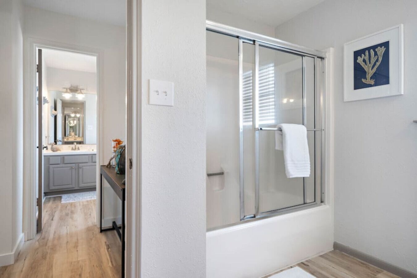 A modern bathroom with a glass shower door, a white towel hanging on the door, and a framed artwork on the wall.