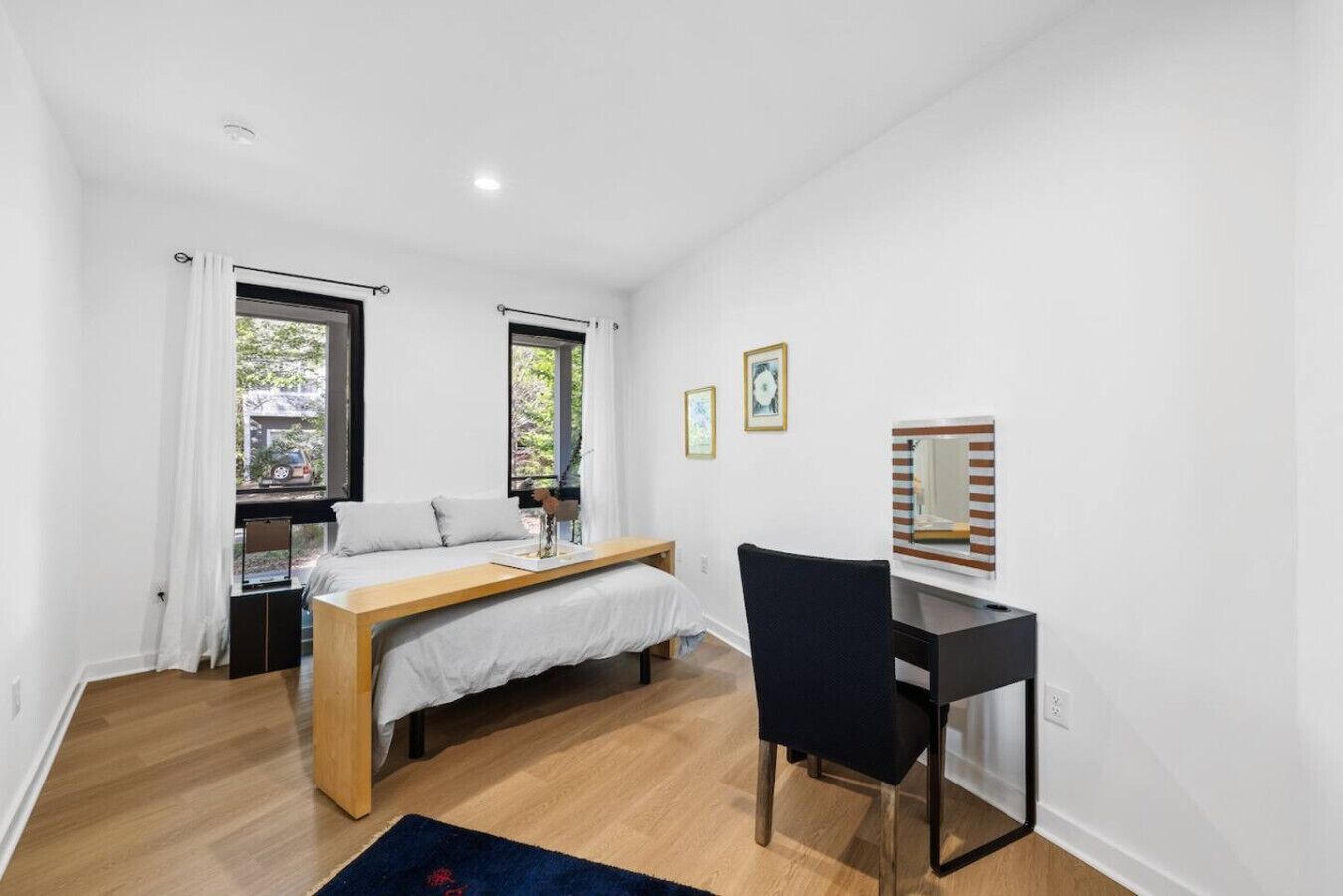 Une chambre avec un lit, un bureau et une chaise. Il y a deux fenêtres avec des rideaux, un miroir et un petit tapis sur le sol.
