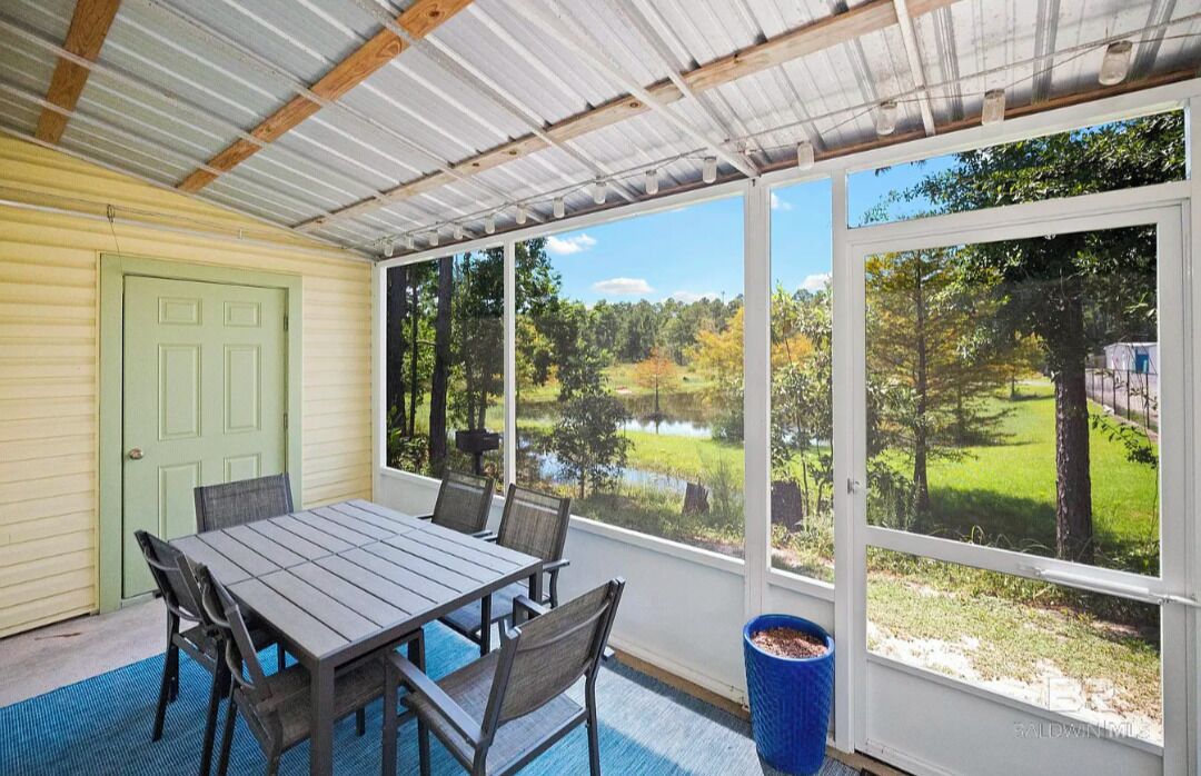 Eine überdachte Terrasse mit Tisch und Stühlen, große Fenster mit Blick auf einen Teich und Bäume.