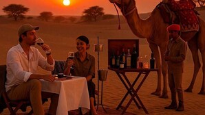 A desert scene with a camel, a table with bottles, and people sitting and standing. - Desert Island Camp & Resort Jaisalmer (Jaisalmer)