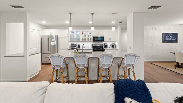A modern kitchen with white cabinetry, a stainless steel refrigerator, a microwave, and a built-in oven. There are three wicker chairs with a blue cushion in front of the kitchen island. - Walk to dinner, near Universal, EV charger, baby/pet gear included, fenced yard (Frisco)