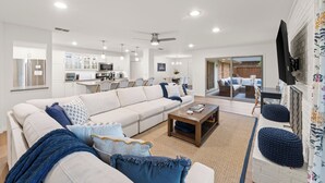 A modern living room with a sofa, coffee table, and ottoman, a kitchen in the background, and a TV mounted on the wall. - Walk to dinner, near Universal, EV charger, baby/pet gear included, fenced yard (Frisco)