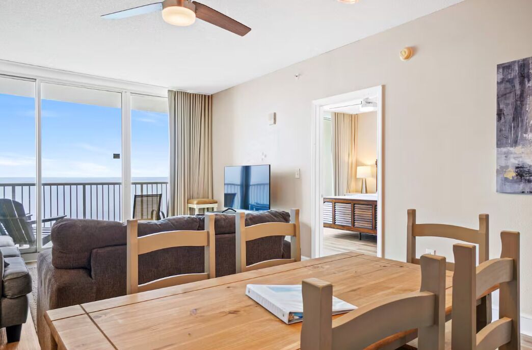 Een kamer met een houten eettafel, stoelen, een televisie en een balkon met uitzicht op zee.