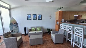 A spacious living area with a sofa, two armchairs, a coffee table, and a bar area with stools. There are framed pictures on the wall, a potted plant, and a surfboard decoration.