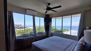 A bedroom with a bed, a ceiling fan, a window seat with cushions, and a view of the ocean.