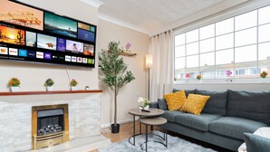 En moderne stue med pejs, en grå sofa med gule puder, et fladskærms-tv monteret på væggen der viser forskellige streamingtjenester, og et stort vindue med udsigt til bygninger.