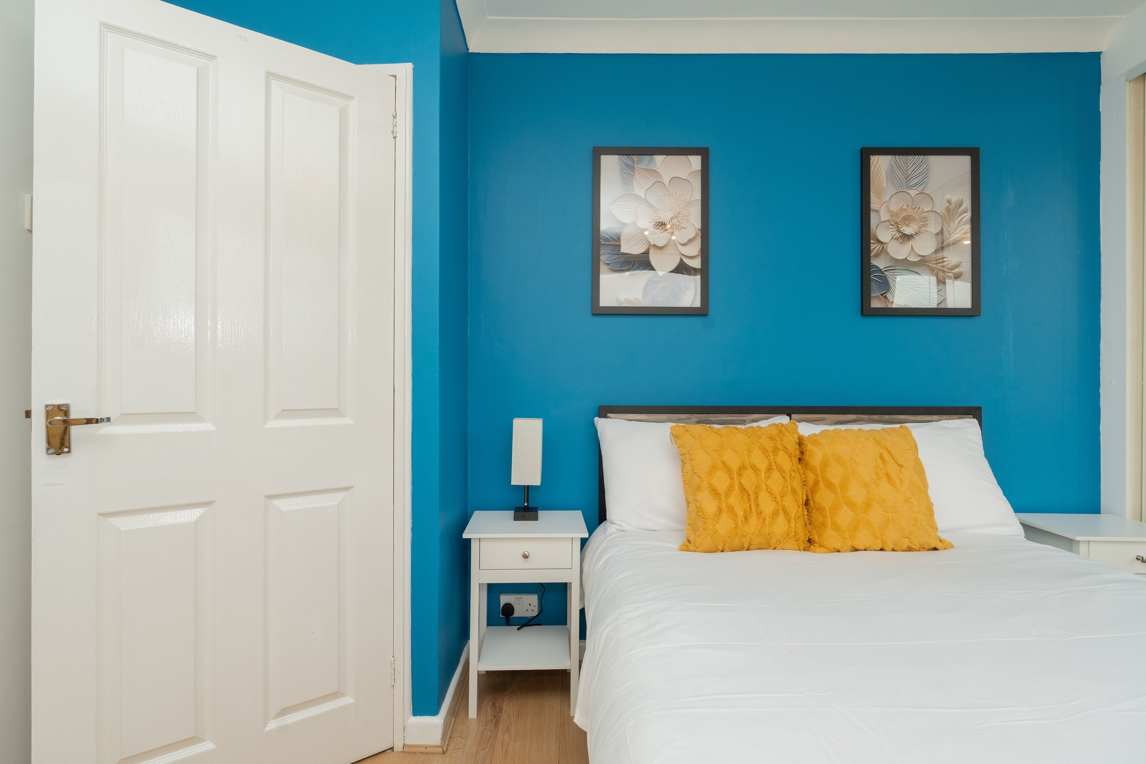 Une chambre avec un mur bleu, un lit blanc et deux tableaux encadrés représentant des motifs floraux.