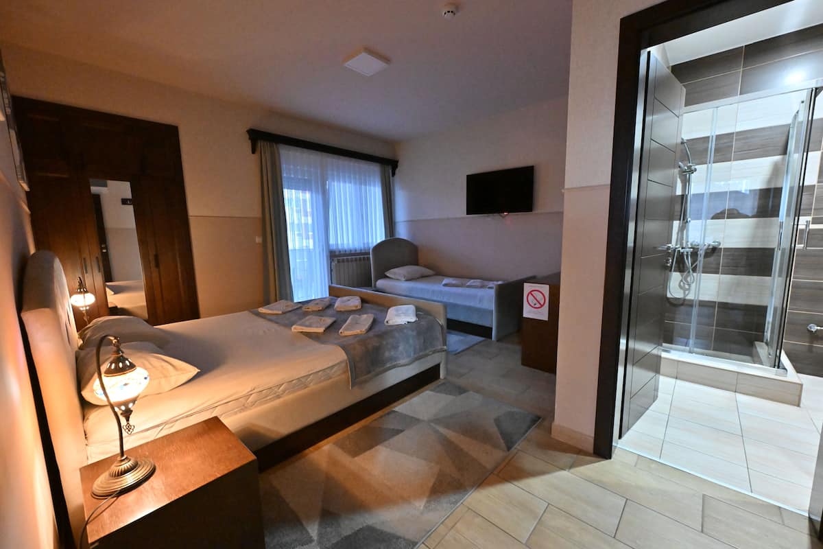 A hotel room with a bed, bedside table, lamp, window with curtains, and a shower area.