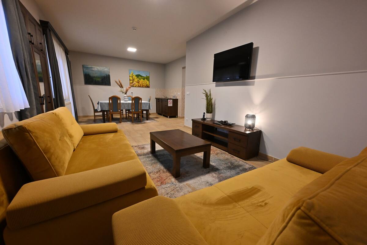 A living room with a yellow sofa set, a coffee table, a TV mounted on the wall, and a dining area with a table and chairs.