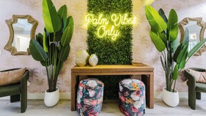 A room with a wall-mounted neon sign reading "Palm Vibes Only," large green plants, a wooden console table with decorative items, and matching upholstered chairs. - Serenity Stays (West Palm Beach)