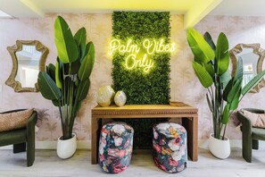 Una habitación con un letrero de neón en la pared que dice "Palm Vibes Only", plantas verdes grandes, una consola de madera con objetos decorativos y sillones a juego tapizados.