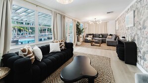A modern living room with a black sofa, grey sofa, and a black coffee table. There is a large window with curtains, a chandelier, and a patterned rug. - Serenity Stays (West Palm Beach)