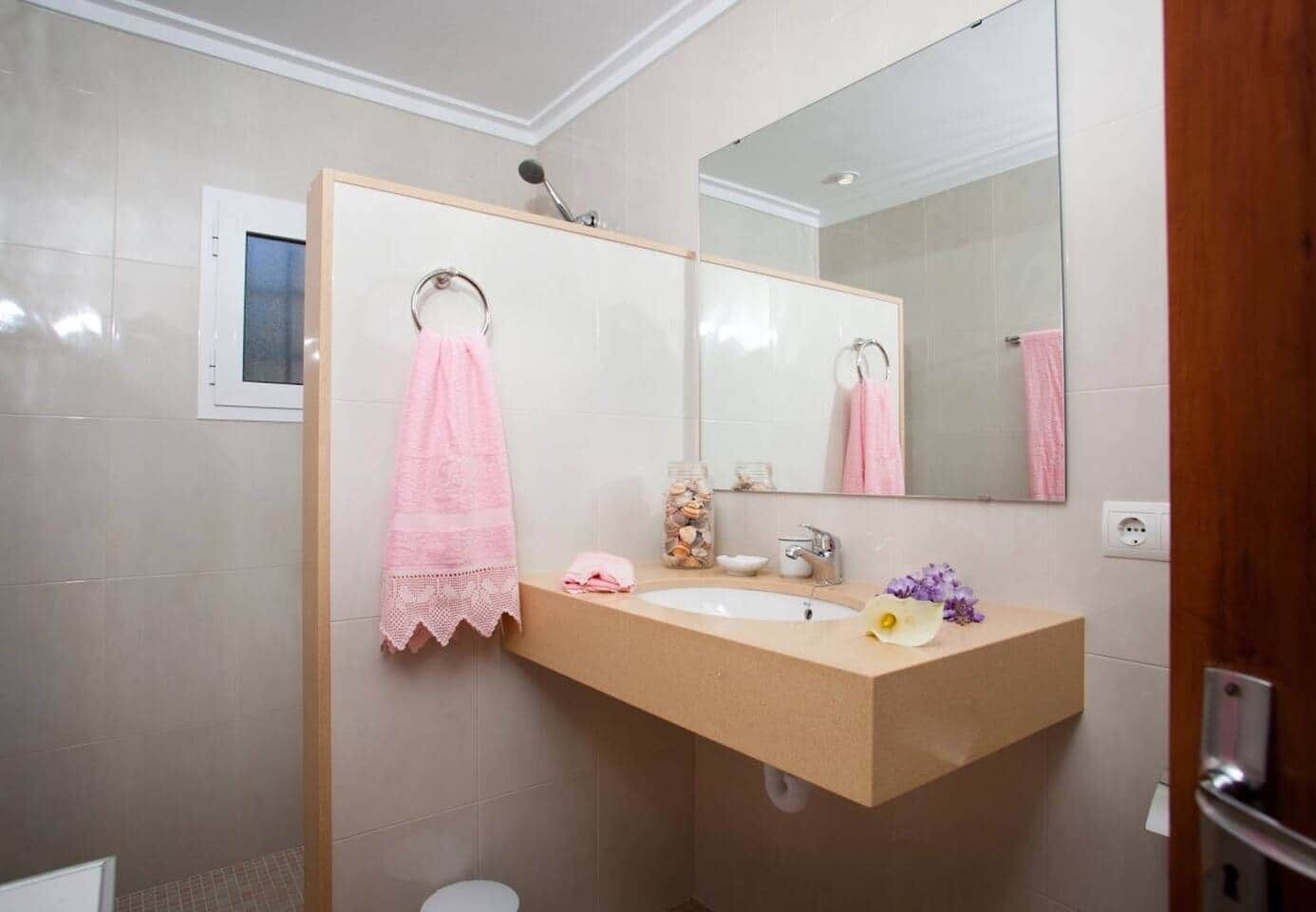 Ein Badezimmer mit einem beige farbenen Waschbecken, einem Spiegel, einem Handtuchhalter mit rosa Handtüchern, einem Glas mit Meeresschnecken und einer Blume auf der Ablagefläche.