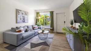 A living room with a grey sofa, a round coffee table, a painting on the wall, a window with curtains, and a door. - Urban Oasis • Modern Comfort in Prime Location (Miami)