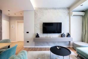 A modern living room with a flat-screen TV mounted on a marble wall, a grey console table with decorative items, a round black coffee table, and a light blue sofa. - Agate Hotel Tirana (Tirana)