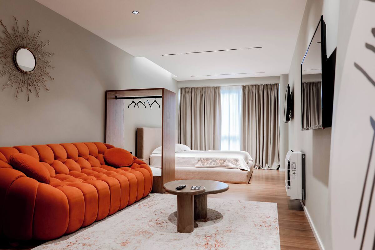 A modern hotel room with a tufted orange sofa, a small round table, a bed with curtains, a mounted TV, and a wall-mounted mirror.
