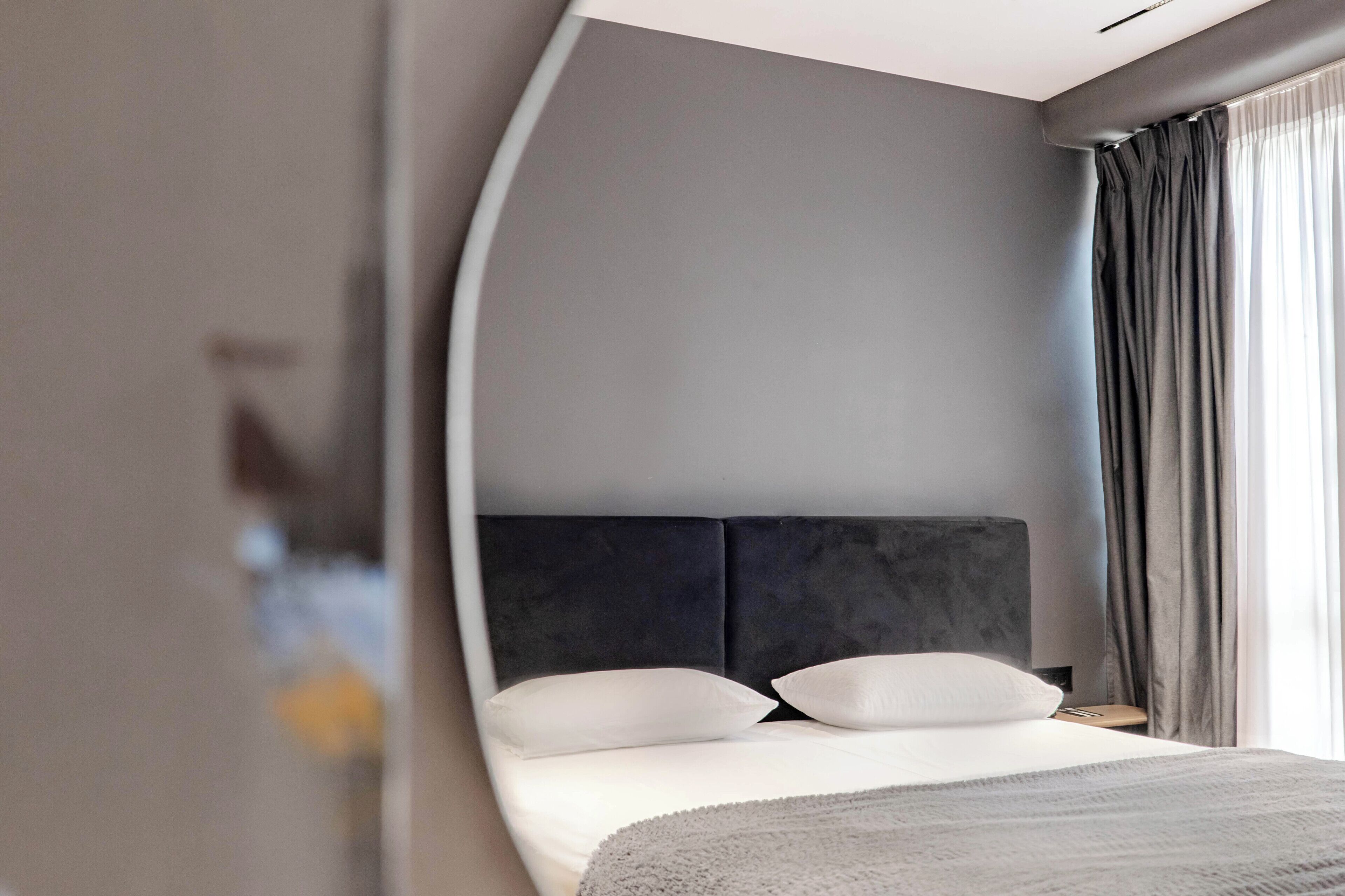 A modern hotel room with a curved wall, a large bed with white linens, a dark headboard, and a view of the outdoors through sheer curtains.