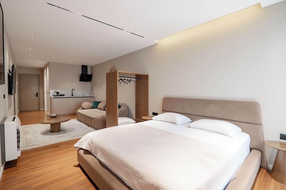 A modern hotel room with a large bed, a small sofa, a dining table, and a kitchenette.