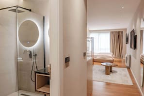 A modern hotel room with a glass-enclosed shower, a round mirror, a small wooden table, and a bed with a white headboard. - Agate Hotel Tirana (Tirana)